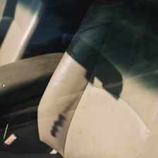 Inside a vehicle, a beige leather car seat with visible wear and creases is depicted. The sunlight casts a distinct shadow across the seat, highlighting its texture. A metallic seatbelt buckle is partially visible in the corner.