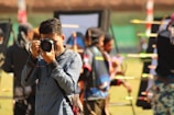 A person in a blue shirt is focusing a camera towards the viewer, with an outdoor archery event happening in the background. Several colorful arrows are visible, stuck in targets. Other people involved in the archery activity are blurred in the background.
