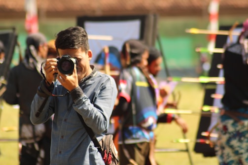 A person in a blue shirt is focusing a camera towards the viewer, with an outdoor archery event happening in the background. Several colorful arrows are visible, stuck in targets. Other people involved in the archery activity are blurred in the background.
