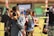 A person in a blue shirt is focusing a camera towards the viewer, with an outdoor archery event happening in the background. Several colorful arrows are visible, stuck in targets. Other people involved in the archery activity are blurred in the background.