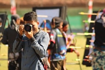 A person in a blue shirt is focusing a camera towards the viewer, with an outdoor archery event happening in the background. Several colorful arrows are visible, stuck in targets. Other people involved in the archery activity are blurred in the background.