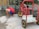 A child wearing a red shirt, blue shorts, and colorful shoes is crouched down near a shallow water feature. Next to the child is an abandoned red metal cart with yellow wheels. The setting appears to be outdoors with a glass barrier and stone paving.