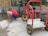A child wearing a red shirt, blue shorts, and colorful shoes is crouched down near a shallow water feature. Next to the child is an abandoned red metal cart with yellow wheels. The setting appears to be outdoors with a glass barrier and stone paving.