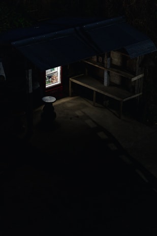 A dimly lit outdoor stall or small structure with a corrugated metal roof and wooden shelves. A refrigerator with a glass door is illuminated, revealing various items inside, possibly food or drinks. The rest of the area is in shadow, with minimal light revealing a small round table.