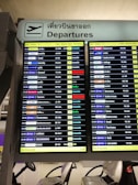 A digital airport departure board showing flight information with details such as time, airlines, destinations, flight numbers, departure gates, and remarks. The sign above is bilingual, displaying 'Departures' in both English and Thai.