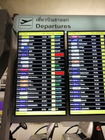 A digital airport departure board showing flight information with details such as time, airlines, destinations, flight numbers, departure gates, and remarks. The sign above is bilingual, displaying 'Departures' in both English and Thai.