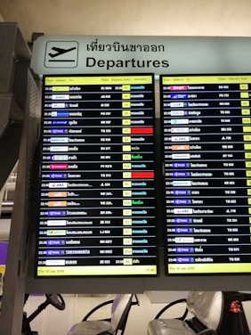 A digital airport departure board showing flight information with details such as time, airlines, destinations, flight numbers, departure gates, and remarks. The sign above is bilingual, displaying 'Departures' in both English and Thai.