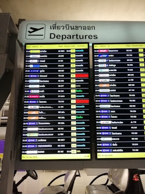 A digital airport departure board showing flight information with details such as time, airlines, destinations, flight numbers, departure gates, and remarks. The sign above is bilingual, displaying 'Departures' in both English and Thai.