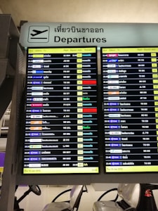 A digital airport departure board showing flight information with details such as time, airlines, destinations, flight numbers, departure gates, and remarks. The sign above is bilingual, displaying 'Departures' in both English and Thai.