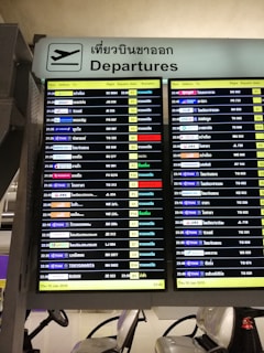 A digital airport departure board showing flight information with details such as time, airlines, destinations, flight numbers, departure gates, and remarks. The sign above is bilingual, displaying 'Departures' in both English and Thai.