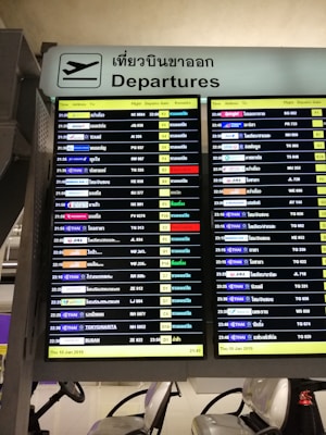 A digital airport departure board showing flight information with details such as time, airlines, destinations, flight numbers, departure gates, and remarks. The sign above is bilingual, displaying 'Departures' in both English and Thai.