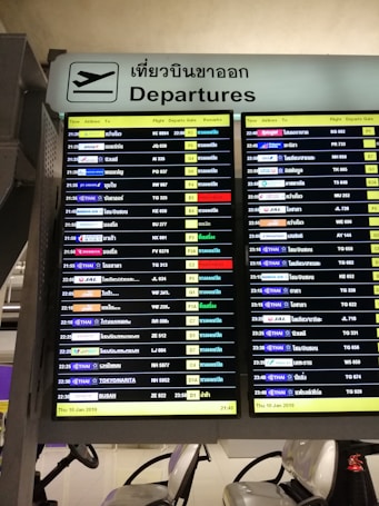 A digital airport departure board showing flight information with details such as time, airlines, destinations, flight numbers, departure gates, and remarks. The sign above is bilingual, displaying 'Departures' in both English and Thai.