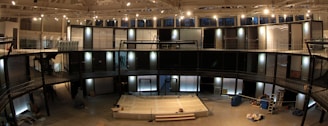 A spacious indoor area with a modern architectural design featuring multiple levels and railings. There is a stage-like platform at the center with objects possibly related to a construction or set-up process scattered around. Large windows adorn the top section, allowing natural light to complement the installed lighting fixtures.