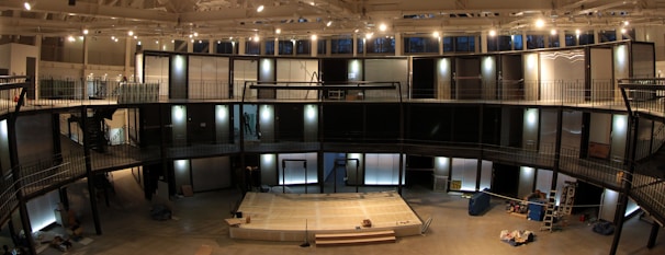 A spacious indoor area with a modern architectural design featuring multiple levels and railings. There is a stage-like platform at the center with objects possibly related to a construction or set-up process scattered around. Large windows adorn the top section, allowing natural light to complement the installed lighting fixtures.