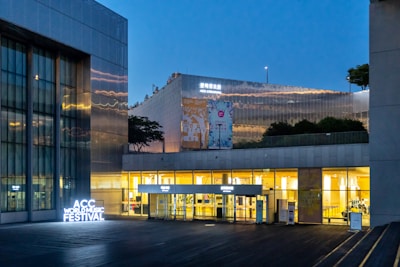 A building with metal and glass architecture illuminated against a twilight sky. Bright signage indicates the ACC World Music Festival, and banners are displayed on the building facade.
