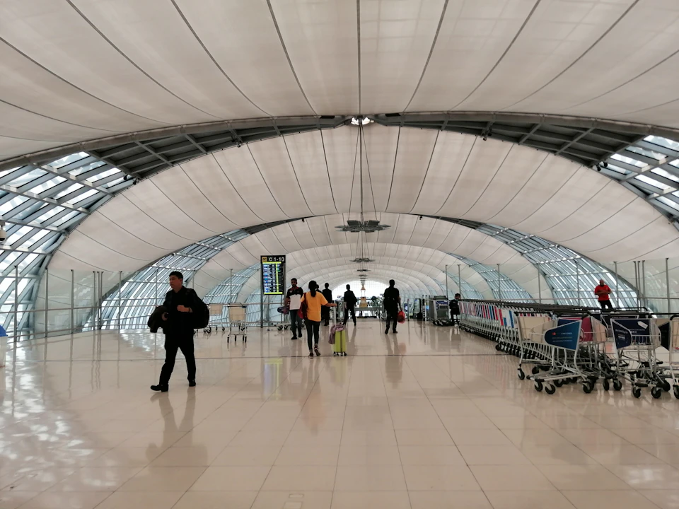 The image depicts a busy airport terminal with a large, curved white ceiling supported by metal beams and covered in a white tarp. The terminal is filled with people walking around, some carrying luggage such as suitcases and handbags. A few individuals are also seen pushing strollers or wheelchairs. In the background, there are signs indicating various locations within the airport, including "Luggage Claim", "Security Checkpoint", and "Ticketing".