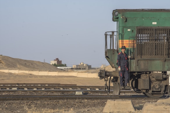 A person in uniform stands on the side of a green train locomotive. The train is situated on railway tracks in a semi-arid landscape, with a barren hill and industrial buildings in the background under a clear blue sky.