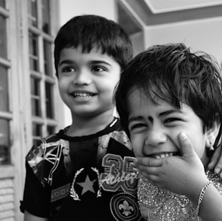 Two cheerful children are standing close to each other, smiling and laughing. One child has their hand covering their mouth in a playful manner, while the other has a wide smile showing their teeth. They are wearing patterned and decorative clothing, and the setting appears to be indoors near a window with wooden framing.