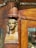 A carved wooden structure features a small, intricate bust of an Egyptian-style figure integrated into the corner of a wooden frame. The wood is polished, showcasing rich brown tones and detailed craftsmanship in both the sculpture and the surrounding woodwork.