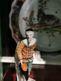 A ceramic figurine depicting a person wearing a detailed costume with a ruffled collar, holding a black hat in one hand. The figure has an intricate design, with an orange jacket, green pants, and fine detailing in the face and clothing. In the background, a decorative plate with floral and landscape designs is visible.