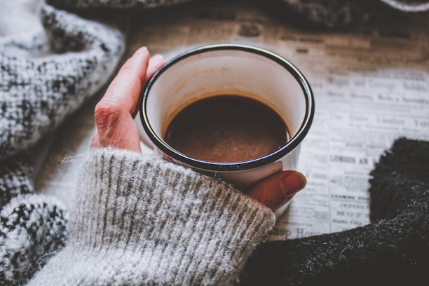 A person wearing a cozy, gray knit sweater holds a cup of coffee, with a newspaper spread out underneath. The scene conveys a sense of warmth and relaxation.