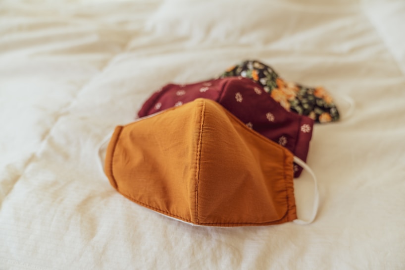 Two fabric face masks are resting on a light-colored bedspread. The mask in the foreground is mustard yellow, while the one in the background has a dark pattern with floral designs.