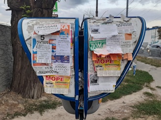 A public bulletin board with a variety of colorful paper advertisements and notices attached, placed outdoors near a tree. The board is surrounded by a pavement and some grass, with vehicles visible in the background driving along a road.