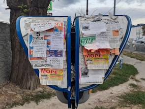 A public bulletin board with a variety of colorful paper advertisements and notices attached, placed outdoors near a tree. The board is surrounded by a pavement and some grass, with vehicles visible in the background driving along a road.