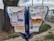 A public bulletin board with a variety of colorful paper advertisements and notices attached, placed outdoors near a tree. The board is surrounded by a pavement and some grass, with vehicles visible in the background driving along a road.