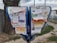 A public bulletin board with a variety of colorful paper advertisements and notices attached, placed outdoors near a tree. The board is surrounded by a pavement and some grass, with vehicles visible in the background driving along a road.