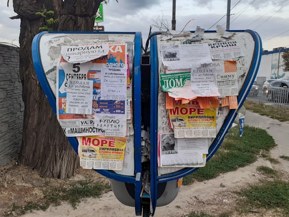 A public bulletin board with a variety of colorful paper advertisements and notices attached, placed outdoors near a tree. The board is surrounded by a pavement and some grass, with vehicles visible in the background driving along a road.