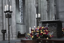 An ornate altar inside a grand cathedral, featuring a decorative metal stand with lit white candles and a vibrant floral arrangement of red, yellow, and blue flowers. The setting showcases large stone pillars and arched windows, creating a solemn and reverent atmosphere.