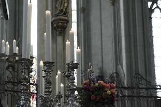 Tall candles positioned on ornate, silver candleholders stand prominently in what appears to be a grand, dimly lit church setting. Stained glass windows and decorative stonework details adorn the background, and a colorful floral arrangement adds vibrant contrast.