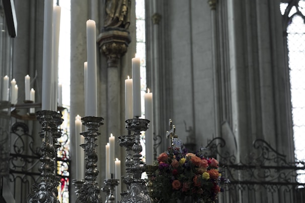Tall candles positioned on ornate, silver candleholders stand prominently in what appears to be a grand, dimly lit church setting. Stained glass windows and decorative stonework details adorn the background, and a colorful floral arrangement adds vibrant contrast.