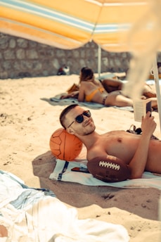 A man is lying on a beach towel under an umbrella, wearing sunglasses and holding a book. Next to him is an orange basketball and a brown football. In the background, another person is lying on a towel wearing a swimsuit.