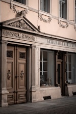 A vintage storefront with detailed architectural elements, including ornate carvings above the double wooden doors and decorative stonework. The windows reflect a classic style with framed glass showcasing an interior display.