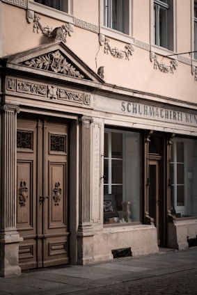 A vintage storefront with detailed architectural elements, including ornate carvings above the double wooden doors and decorative stonework. The windows reflect a classic style with framed glass showcasing an interior display.