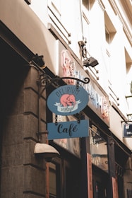 A quaint café sign hangs outside an establishment on a stone building. The sign features a blue background with a pink elephant and the words 'Delirium Village'. The architecture reflects an urban setting with large windows and subtle lighting. The street sign suggests a cozy and inviting atmosphere.
