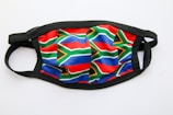 A face mask featuring the pattern of the South African flag. It has detailed red, blue, green, yellow, black, and white elements. The background is plain white and the mask edge is black in color.