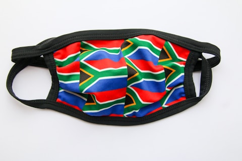 A face mask featuring the pattern of the South African flag. It has detailed red, blue, green, yellow, black, and white elements. The background is plain white and the mask edge is black in color.