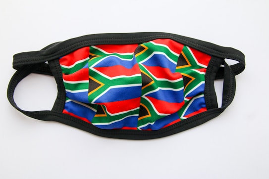 A face mask featuring the pattern of the South African flag. It has detailed red, blue, green, yellow, black, and white elements. The background is plain white and the mask edge is black in color.
