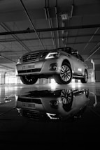 A silver SUV is parked indoors with its headlights on, casting a clear reflection on the polished floor. The surrounding environment appears to be a spacious parking garage with visible ceiling pipes and structures.