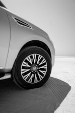A detailed shot of a car's front section, focusing on a modern alloy wheel and tire with intricate patterns. The car is positioned on a sandy surface with noticeable tire tracks, and the scene is lit brightly, suggesting a clear, sunny day.