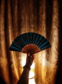 A hand holding an open, blue folding fan against a curtain illuminated by a lamp in the background. The curtain has an ornate pattern, creating a warm and cozy ambiance with its golden hue.