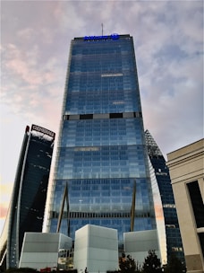 A tall, modern skyscraper with a sleek glass facade stands prominently against a backdrop of a partly cloudy sky. The skyscraper features a blue logo at the top and is flanked by other architecturally distinct towers. The surrounding environment includes smaller structures and some greenery at the base.