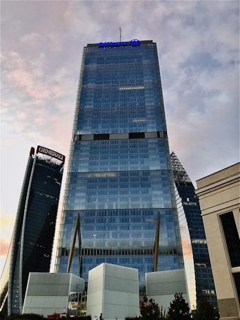 A tall, modern skyscraper with a sleek glass facade stands prominently against a backdrop of a partly cloudy sky. The skyscraper features a blue logo at the top and is flanked by other architecturally distinct towers. The surrounding environment includes smaller structures and some greenery at the base.