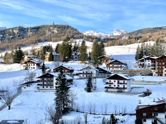 A quaint alpine village nestled in a snowy landscape, featuring a cluster of wooden chalets surrounded by snow-covered trees and hills. The background showcases a range of mountains under a clear blue sky.