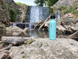 A turquoise water bottle stands on a large rock in front of a scenic waterfall surrounded by rocks and lush greenery. The cascading water creates a serene and natural backdrop. The area is filled with rocks and logs, blending into a tranquil natural environment.