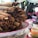 A large stack of crispy rolled tacos, also known as taquitos, is piled on a decorative plate. The plate is on a table outside with blurred figures sitting behind it, likely in a market or fair setting. Various toppings and sauces in bowls or containers are also on the table.