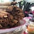 A large stack of crispy rolled tacos, also known as taquitos, is piled on a decorative plate. The plate is on a table outside with blurred figures sitting behind it, likely in a market or fair setting. Various toppings and sauces in bowls or containers are also on the table.